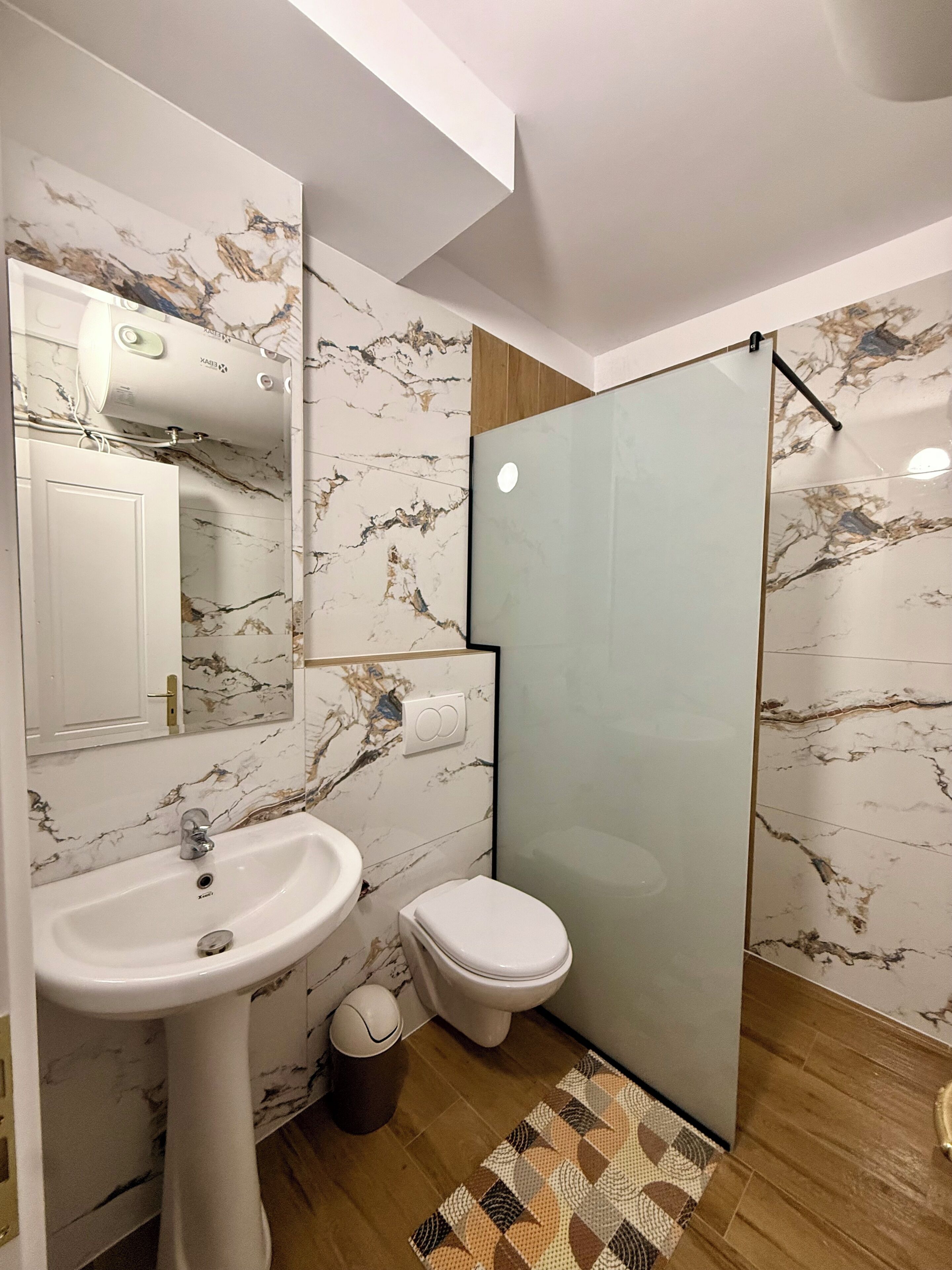 A bathroom with a marble backsplash, a sink, a toilet, and a glass shower enclosure.