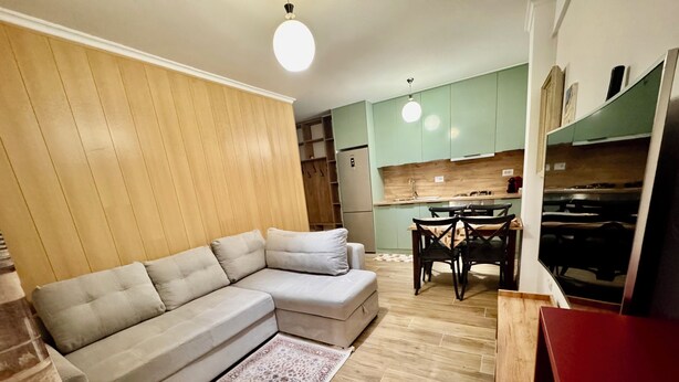 A living room with a sectional sofa, a dining table with chairs, a kitchen area with cabinets, and a red cabinet. - Aisha Apartment (Berat)