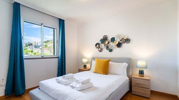 Un lit bien fait, avec du linge de lit blanc et un coussin jaune, deux tables de chevet avec des lampes, un élément décoratif fixé au mur et une fenêtre avec des rideaux bleus.