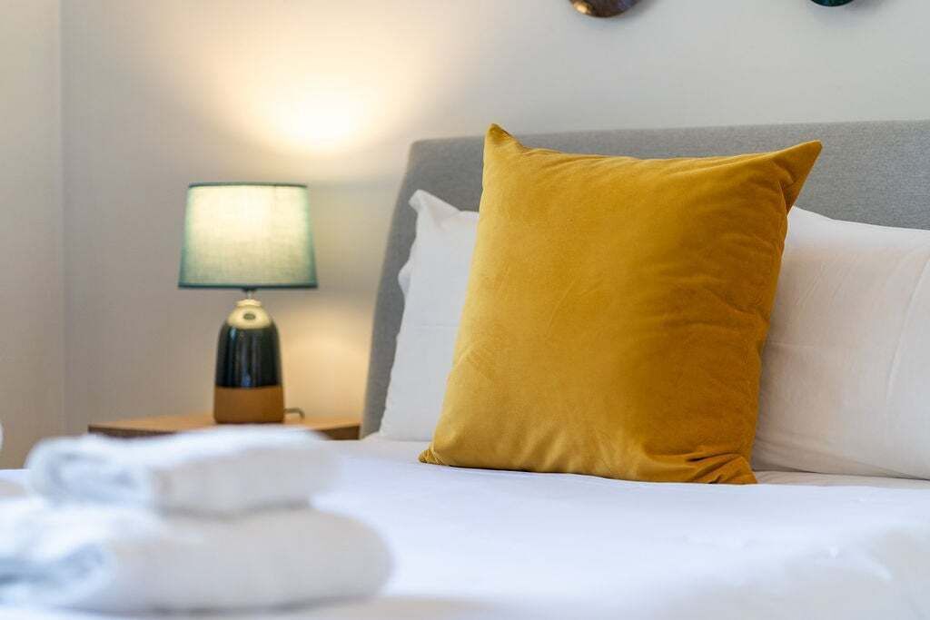 Un lit bien fait, avec une literie blanche, un seul oreiller de couleur moutarde et une lampe de chevet avec un abat-jour vert.