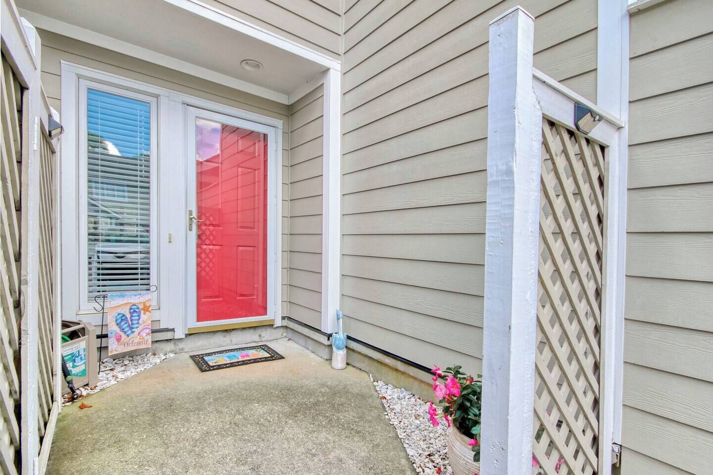 A house entrance with a red door, a welcome mat, and a small garden area with flowers.