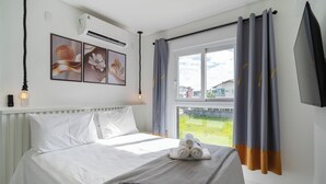 A hotel room with a bed, a window with curtains, a wall-mounted air conditioner, and a wall with framed pictures. - Apts in a Condominium w/ Pool Just 500 m fr Ingleses Beach - Mncs (Florianópolis)