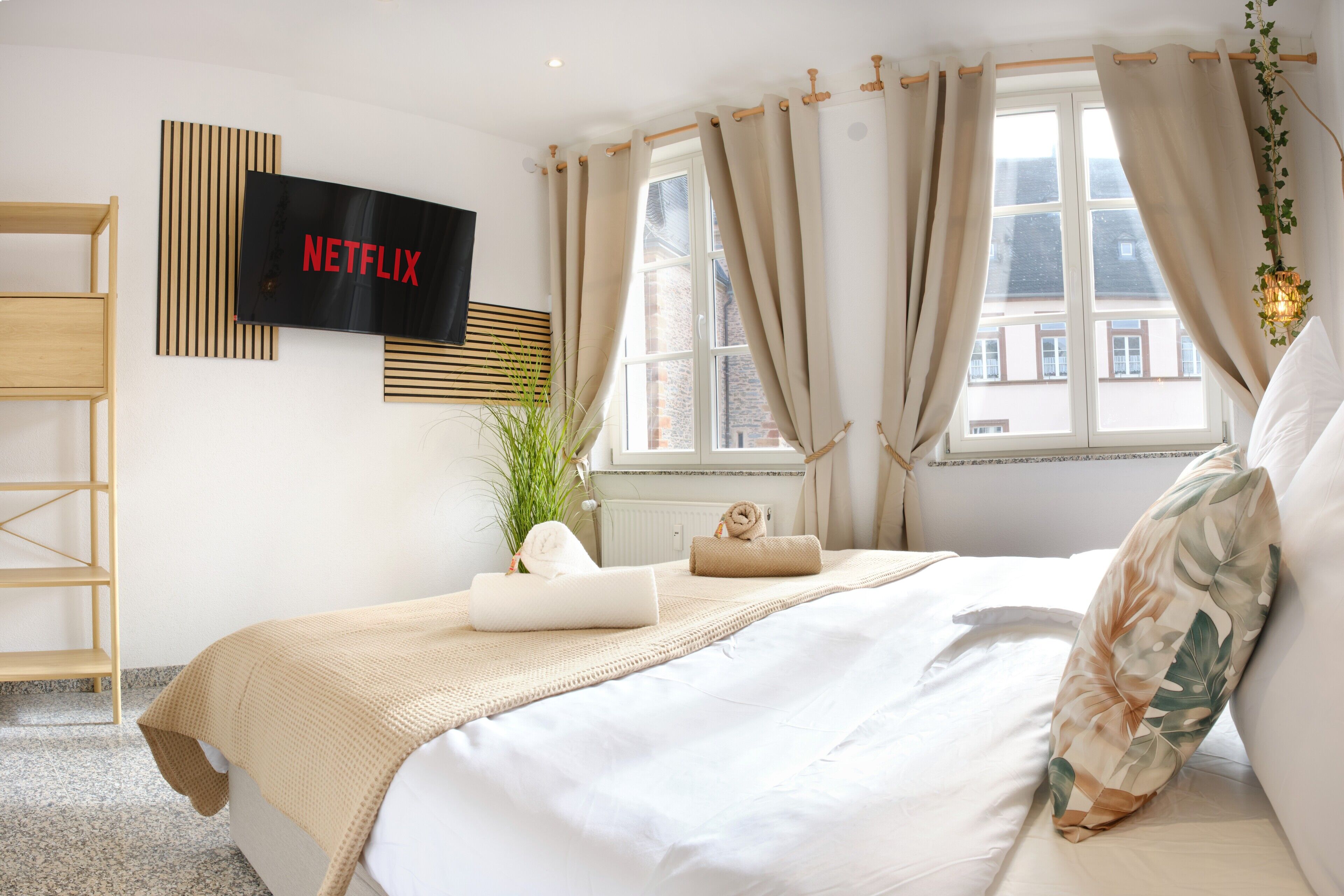 A bedroom with a bed, a TV mounted on the wall, a window with curtains, and a plant.