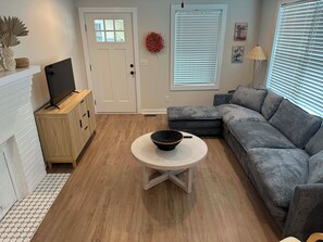 Un salon avec un canapé gris, une table basse ronde, un téléviseur posé sur un meuble en bois, une porte blanche et une fenêtre avec des stores.