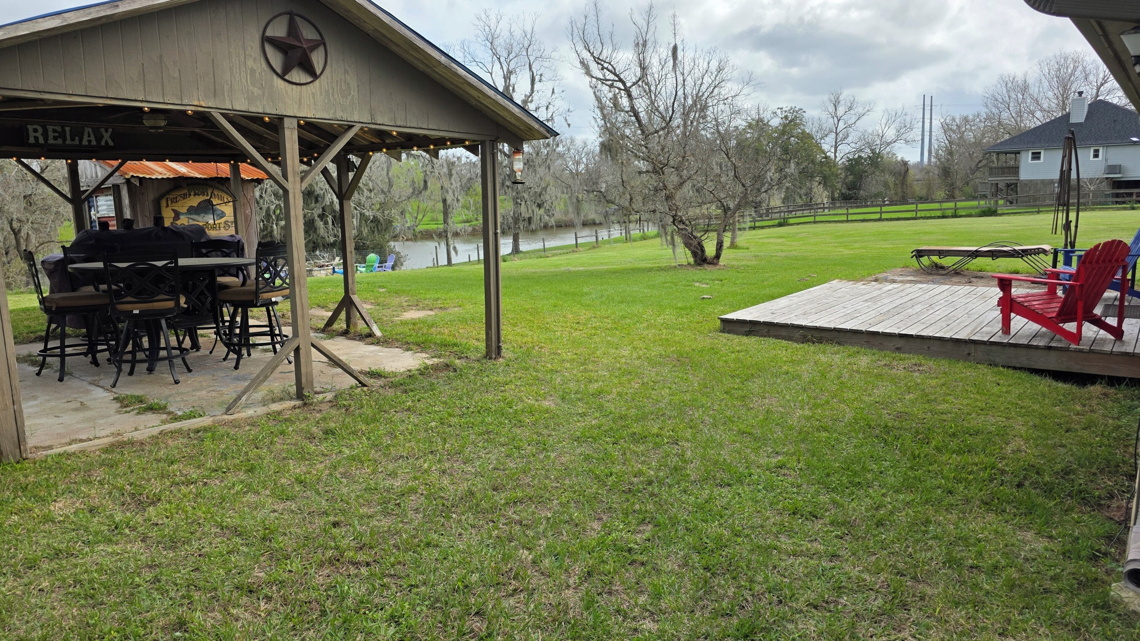 A covered outdoor seating area with a grill, a wooden deck with a red chair, and a grassy yard with a lake in the background.