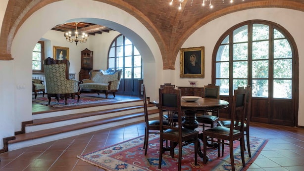 A dining area with a round table, chairs, and a chandelier. There are arched windows and a staircase leading to another room. - Villa nestled in the vineyards of chianti with private pool (Pieve A Presciano)