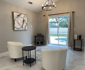 A living room with two chairs, a small table, a chandelier, a bar area, and a view of a swimming pool. - Pool, Park, & Ramp! Book today! (Port St. Lucie)