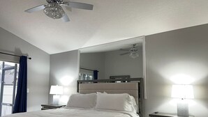 A hotel room with a bed, bedside lamps, a ceiling fan, a large mirror, and a window with curtains. - Pool, Park, & Ramp! Book today! (Port St. Lucie)