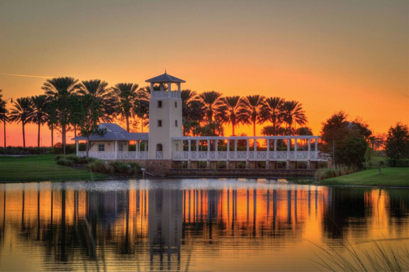 Un bâtiment situé au bord de l’eau, doté d’une tour, entouré de palmiers et surplombant un lac paisible où se reflète le coucher de soleil.