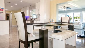 Uma sala de estar moderna com uma mesa de centro de vidro, sofás brancos e uma área de cozinha ao fundo.