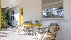 Eine Veranda mit zwei Korbstühlen und einem kleinen runden Tisch, ein Fenster mit Rollläden und eine Tür mit gelbem Rahmen.