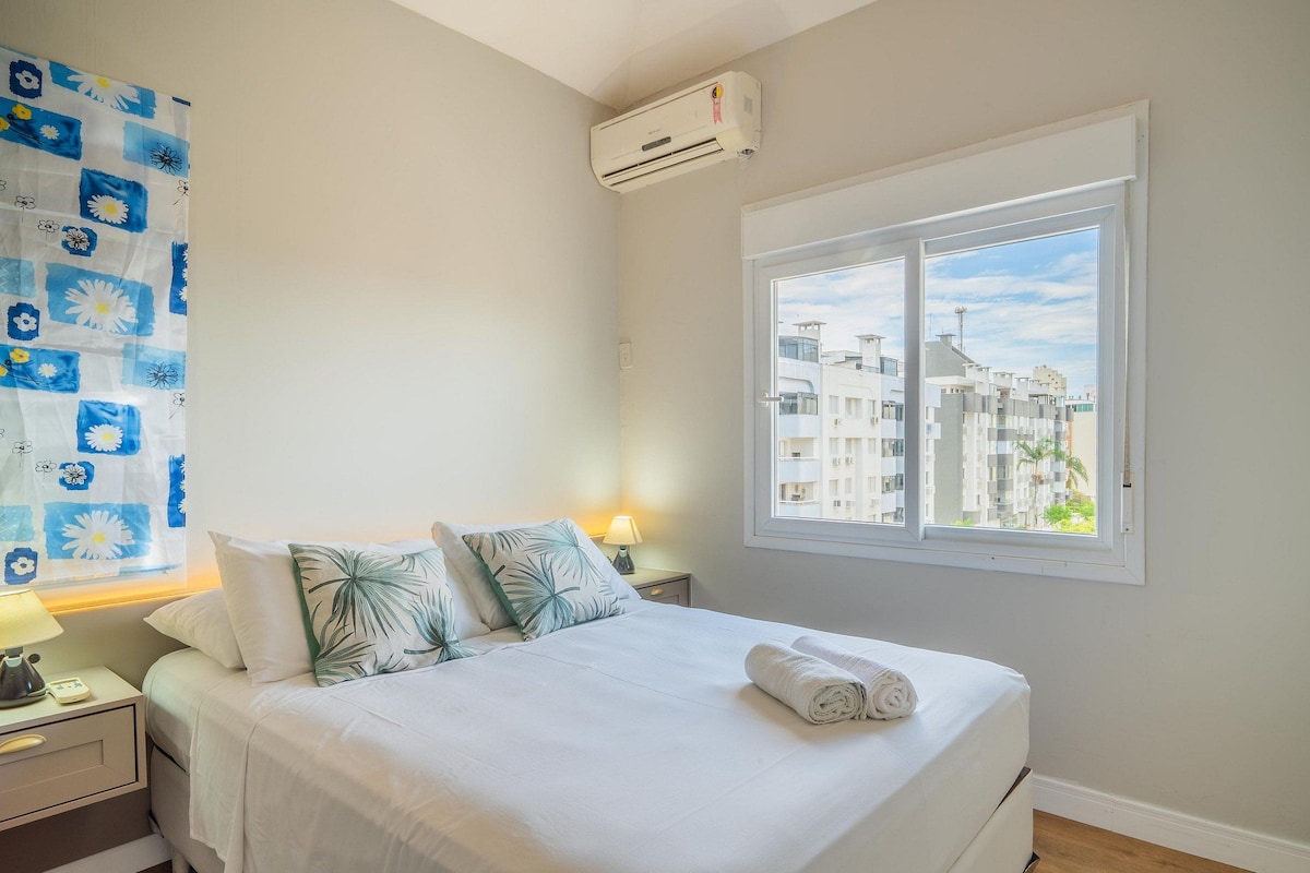 Ein Hotelzimmer mit Bett, Nachttisch, Lampe, Klimaanlage und Fenster mit Blick auf die Stadt.