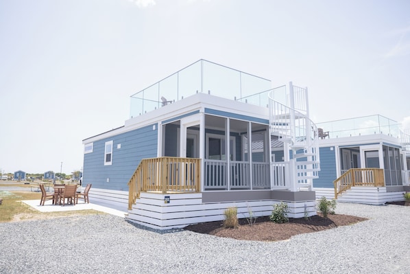A modern, two-story house with a balcony, a porch, and a small outdoor seating area. - Chincoteague Bay RV Resort & Cottages (Chincoteague)