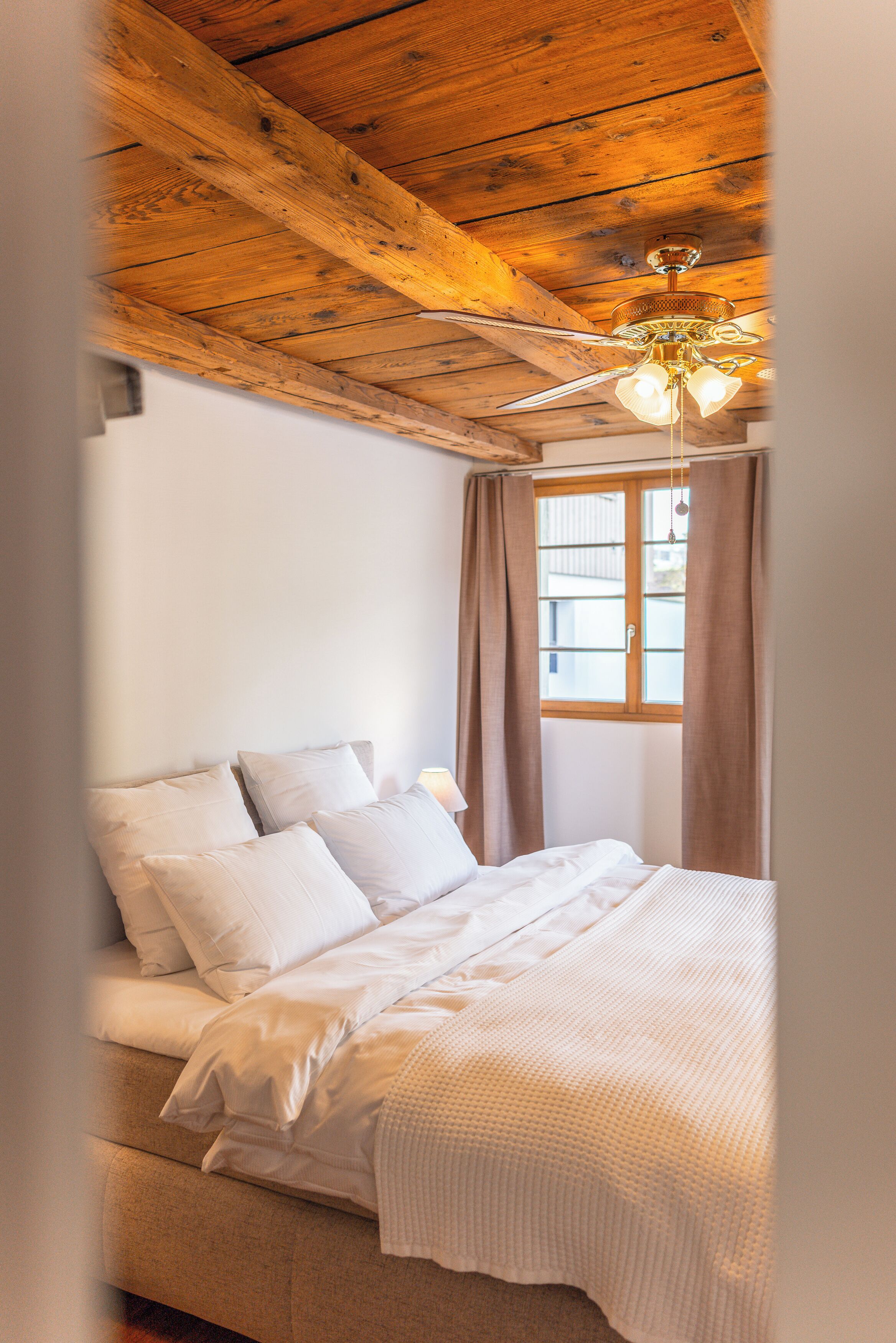 Ein Schlafzimmer mit Holzboden, einem Bett mit weißer Bettwäsche, einem Fenster mit Vorhängen und einem Deckenventilator.