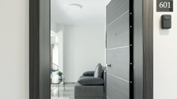 Un interior moderno con una puerta de madera oscura, el número de habitación 601 visible y una vista parcial de una sala de estar con un sofá y una mesita.