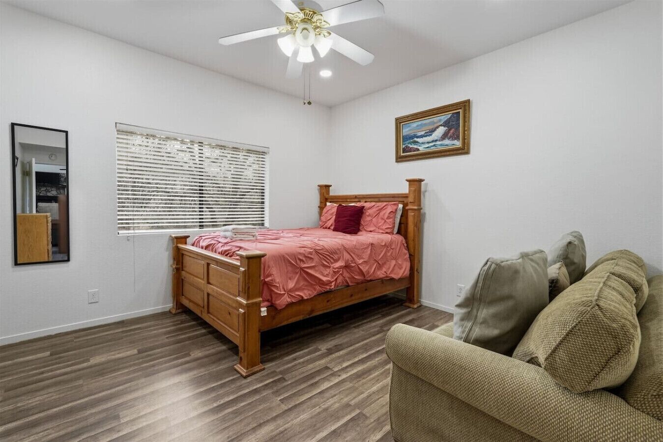 A bedroom with a wooden bed, a ceiling fan, a window with blinds, a framed picture on the wall, and a chair.