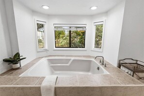 Ein Badezimmer mit einer großen Badewanne, weißen Wänden und einem Fenster mit Blick auf Grünflächen.