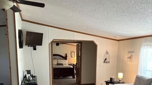 A spacious living area with a wooden floor, a grey sofa, a ceiling fan, a flat-screen TV, and a bed in the background.
