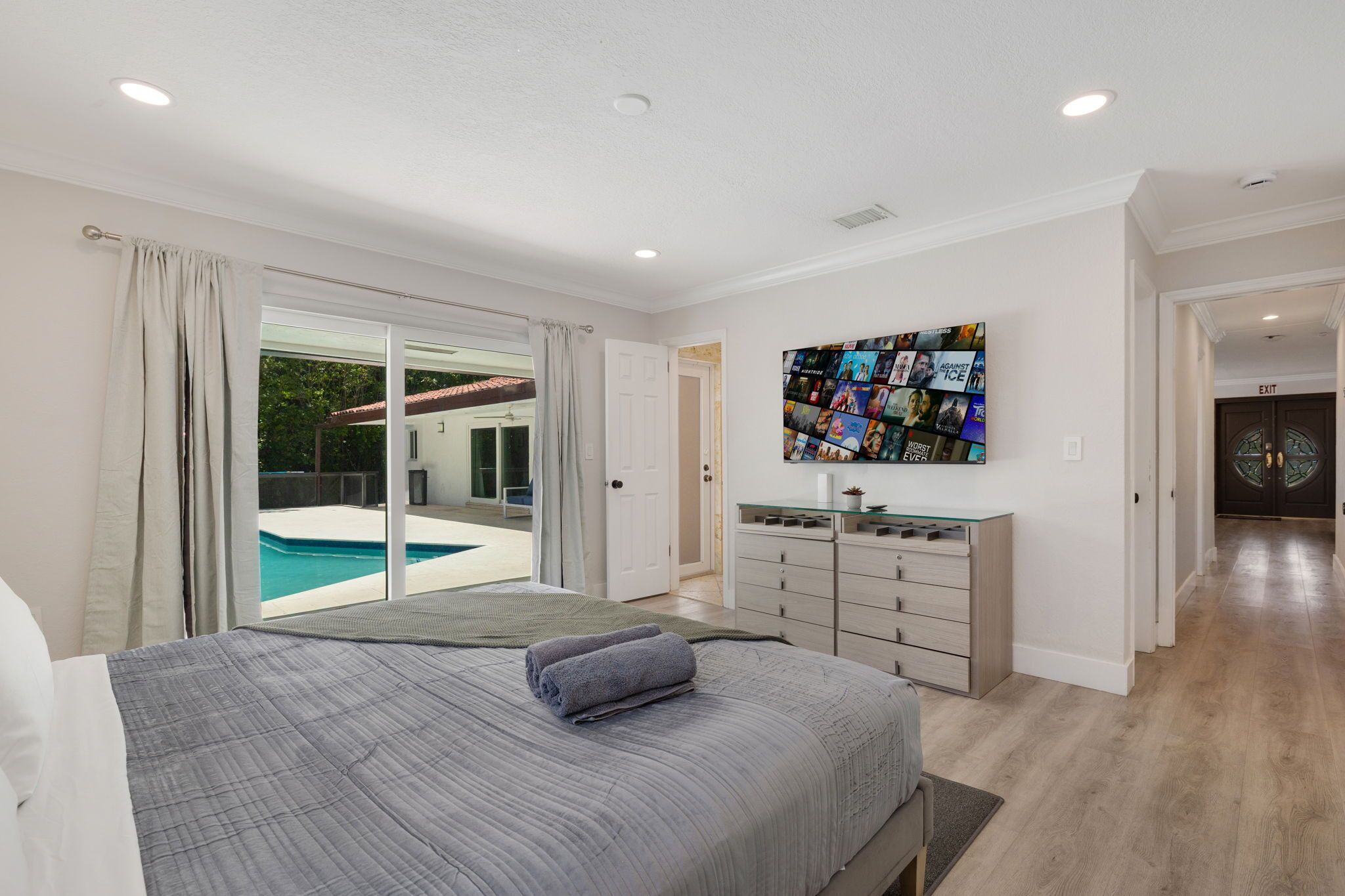 A bedroom with a large bed, a TV mounted on the wall, and a sliding glass door leading to a pool.