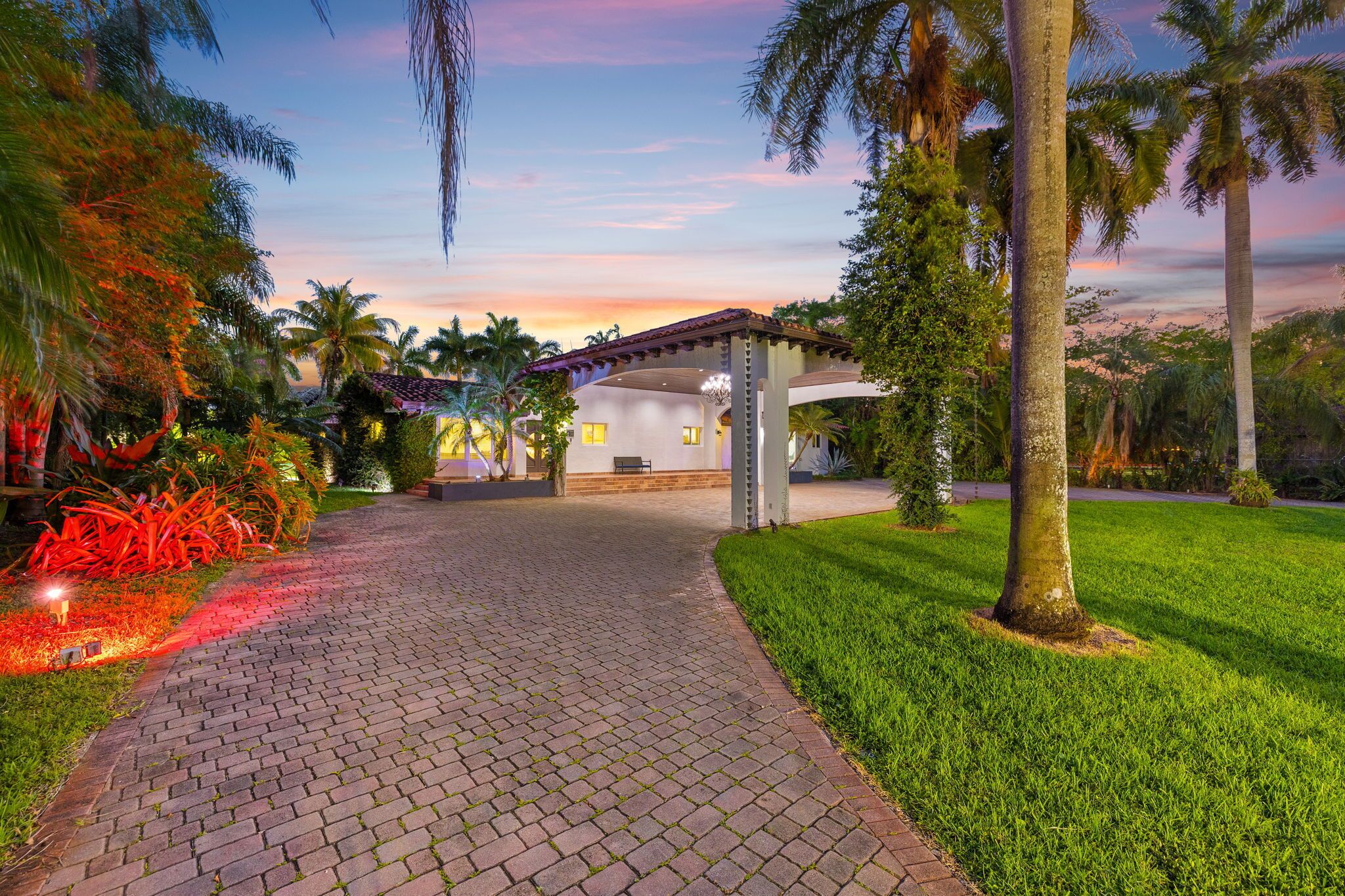 A paved pathway leading to a building with a covered patio area, surrounded by lush greenery and palm trees.