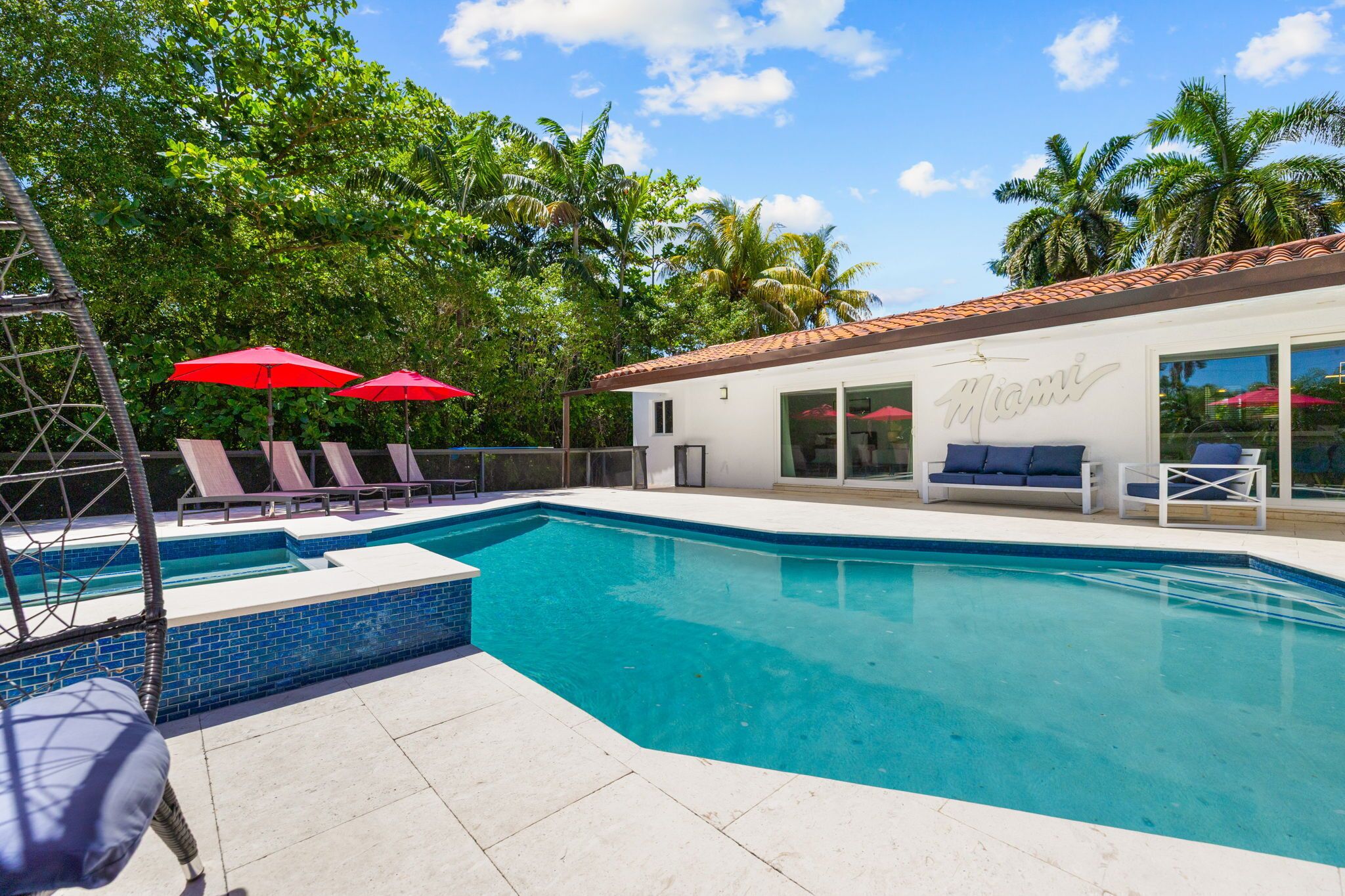 A swimming pool with lounge chairs and umbrellas, a white building with 'Miami' on the side, and palm trees in the background.