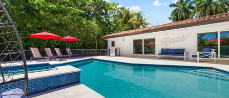 A swimming pool with lounge chairs and umbrellas, a white building with 'Miami' on the side, and palm trees in the background.