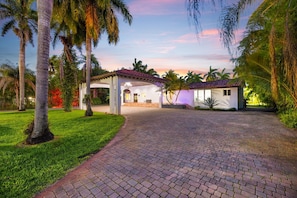 A covered outdoor area with a paved walkway leading to a building, surrounded by palm trees and greenery.