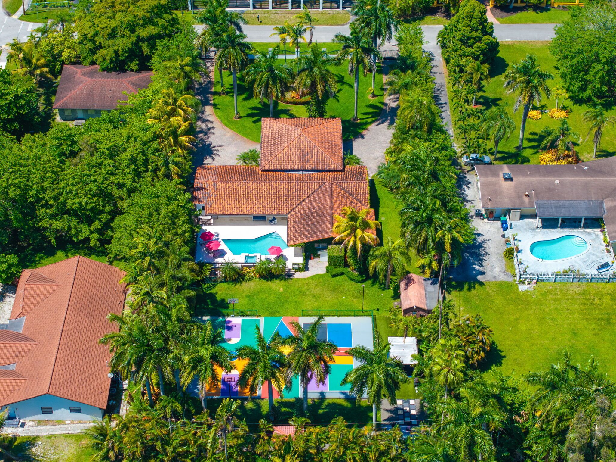 An aerial view of a residential area with a swimming pool, a tennis court, and a large house with a red-tiled roof.