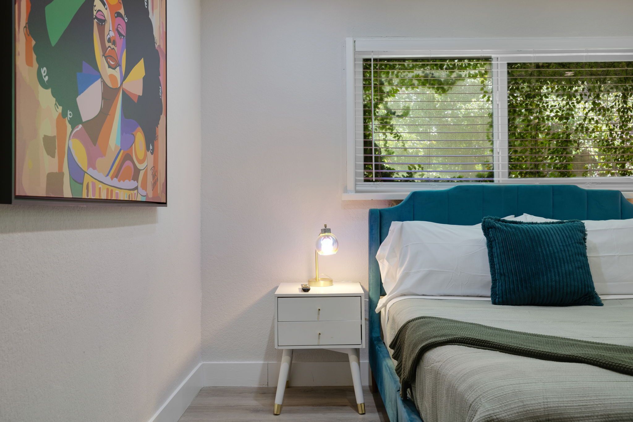 A bedroom with a bed, a nightstand with a lamp, a window with blinds, and a painting on the wall.