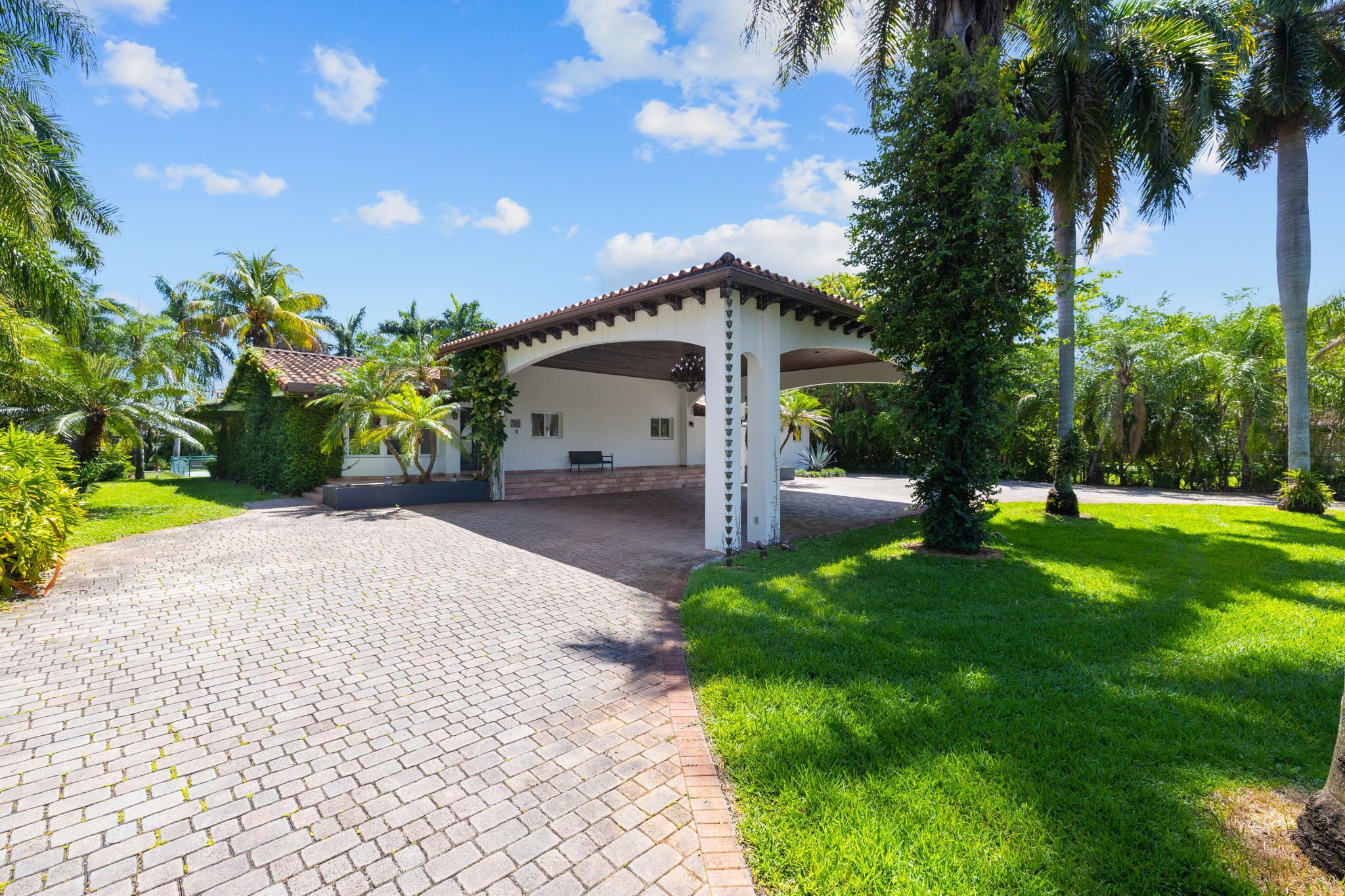 A covered outdoor area with a paved walkway leading to a building, surrounded by lush greenery and palm trees.