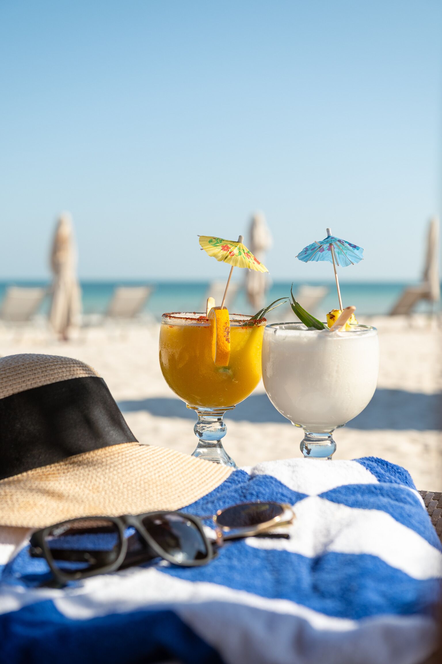 Two drinks on a beach towel, a hat, and sunglasses.