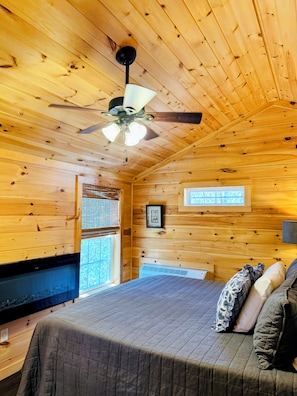 Een slaapkamer in een houten blokhut met een bed, een plafondventilator, een raam met zonwering en een ingelijst beeld aan de muur.