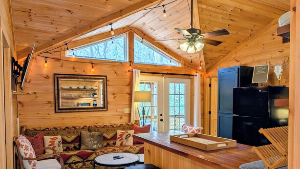 A cozy cabin interior with a wooden ceiling, a sofa with patterned cushions, a small dining table, a bar area, and a black refrigerator. - Cozy Cabin Near Lake Burton Clayton - Dog Friendly! (Clarkesville)