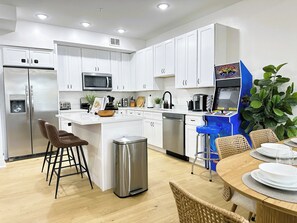 Een moderne keuken met witte kasten, een roestvrijstalen koelkast, een magnetron en een koffiezetapparaat. Er is een eethoek met een houten tafel en rieten stoelen.