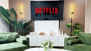 Un soggiorno con divani verdi, un tavolino da caffè bianco, una TV con scritto 'NETFLIX' e una parete in legno con listelli verticali.