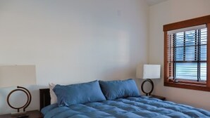 A bedroom with a bed, two bedside tables, a window with blinds, and a wall-mounted lamp. - Inn at Whittier (Whittier)
