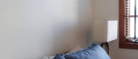 A room with a bed, a nightstand, a window with blinds, and a wall-mounted lamp.