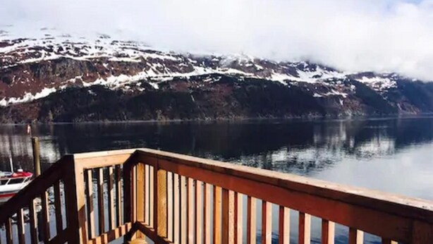 A wooden deck with a railing, leading to a calm lake with snow-capped mountains in the background. - Inn at Whittier (Whittier)