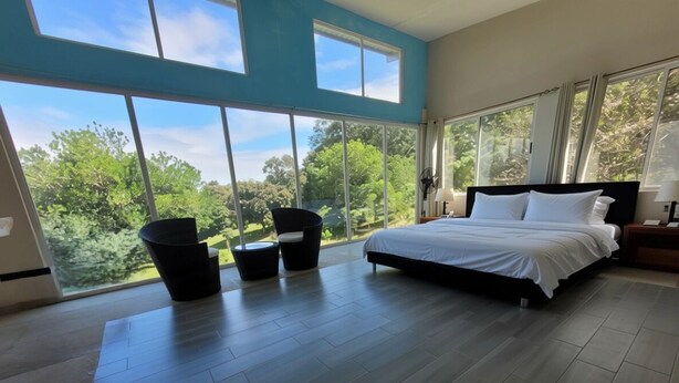 Ein Schlafzimmer mit einem großen Bett, Blick auf Bäume durch große Fenster und einer Sitzecke mit zwei Stühlen.