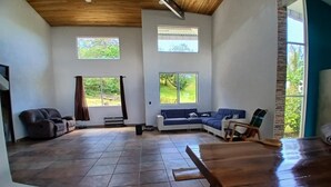 Ein großzügiger Wohnbereich mit Holzbalkendecke, großen Fenstern, einer Sofagarnitur und einem Esstisch.
