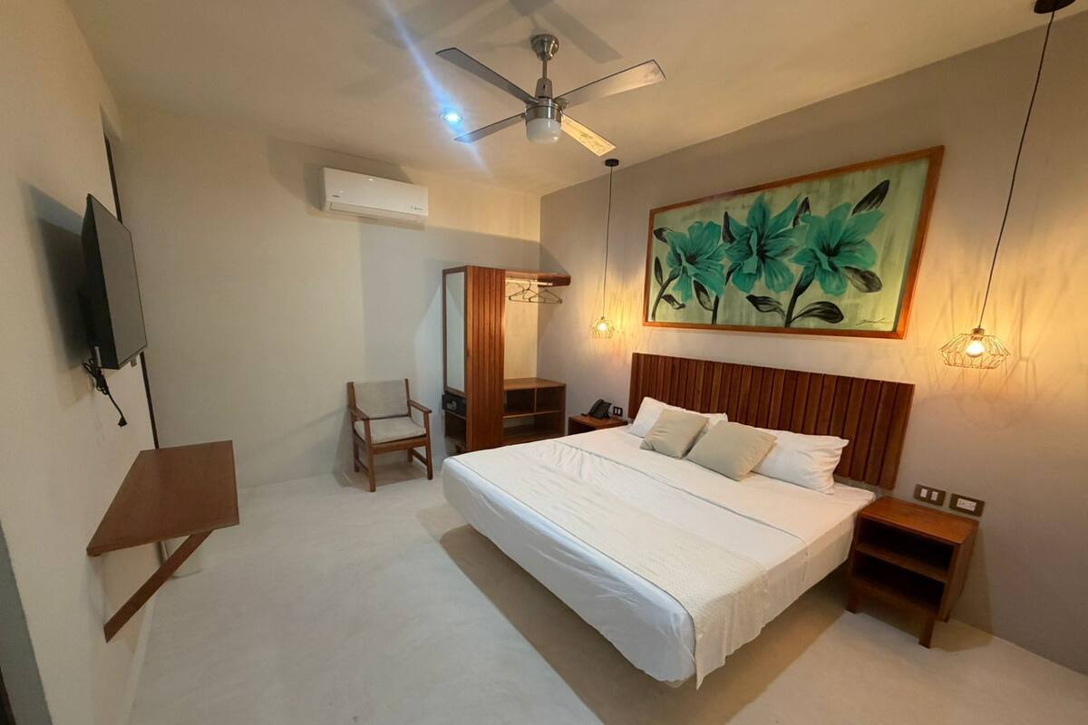 A bedroom with a bed, a chair, a desk, a wardrobe, a ceiling fan, and a painting.