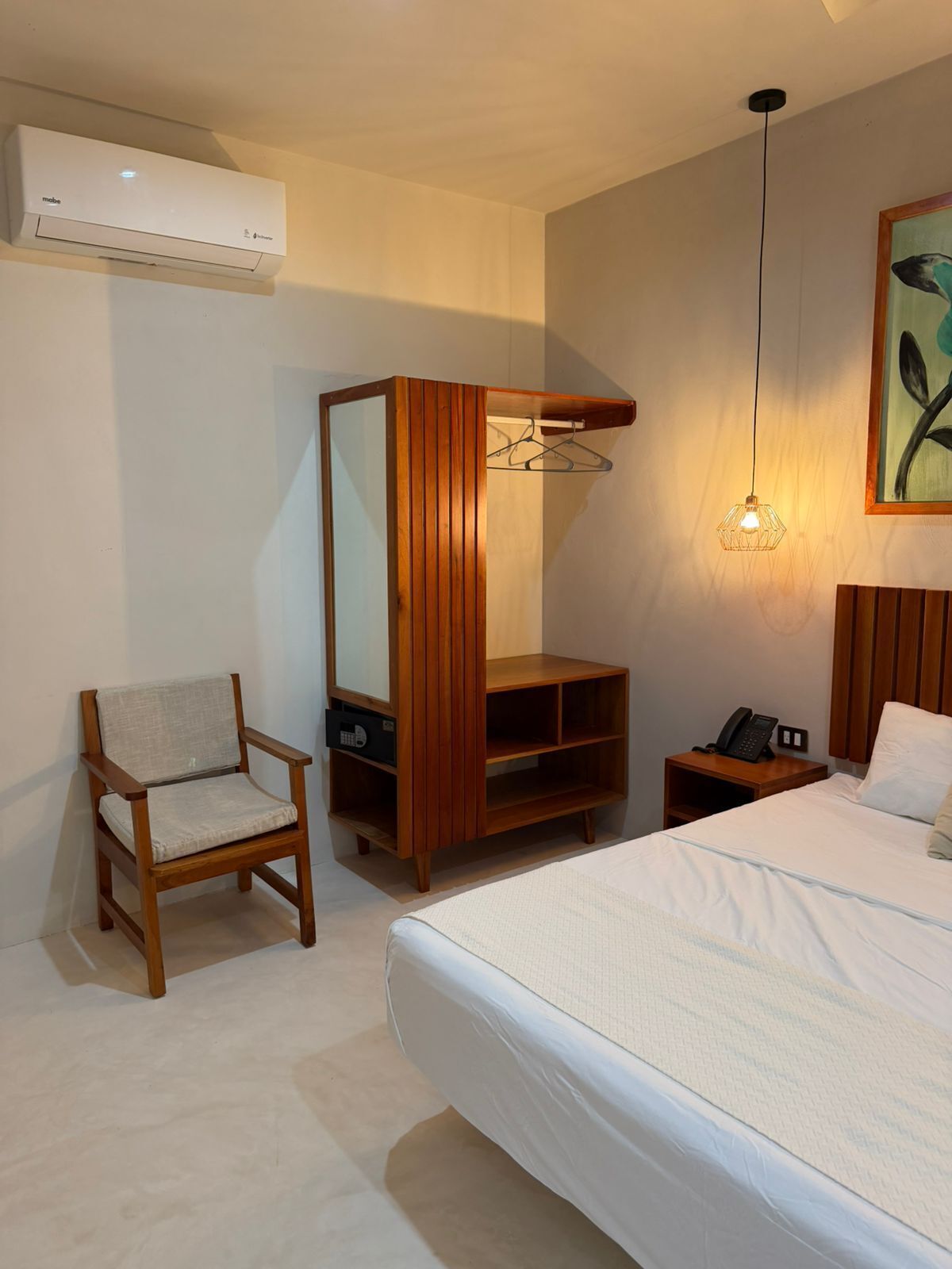 A hotel room with a bed, a wooden chair, a wooden wardrobe with a mirror, a bedside table with a phone, and a wall-mounted air conditioner.
