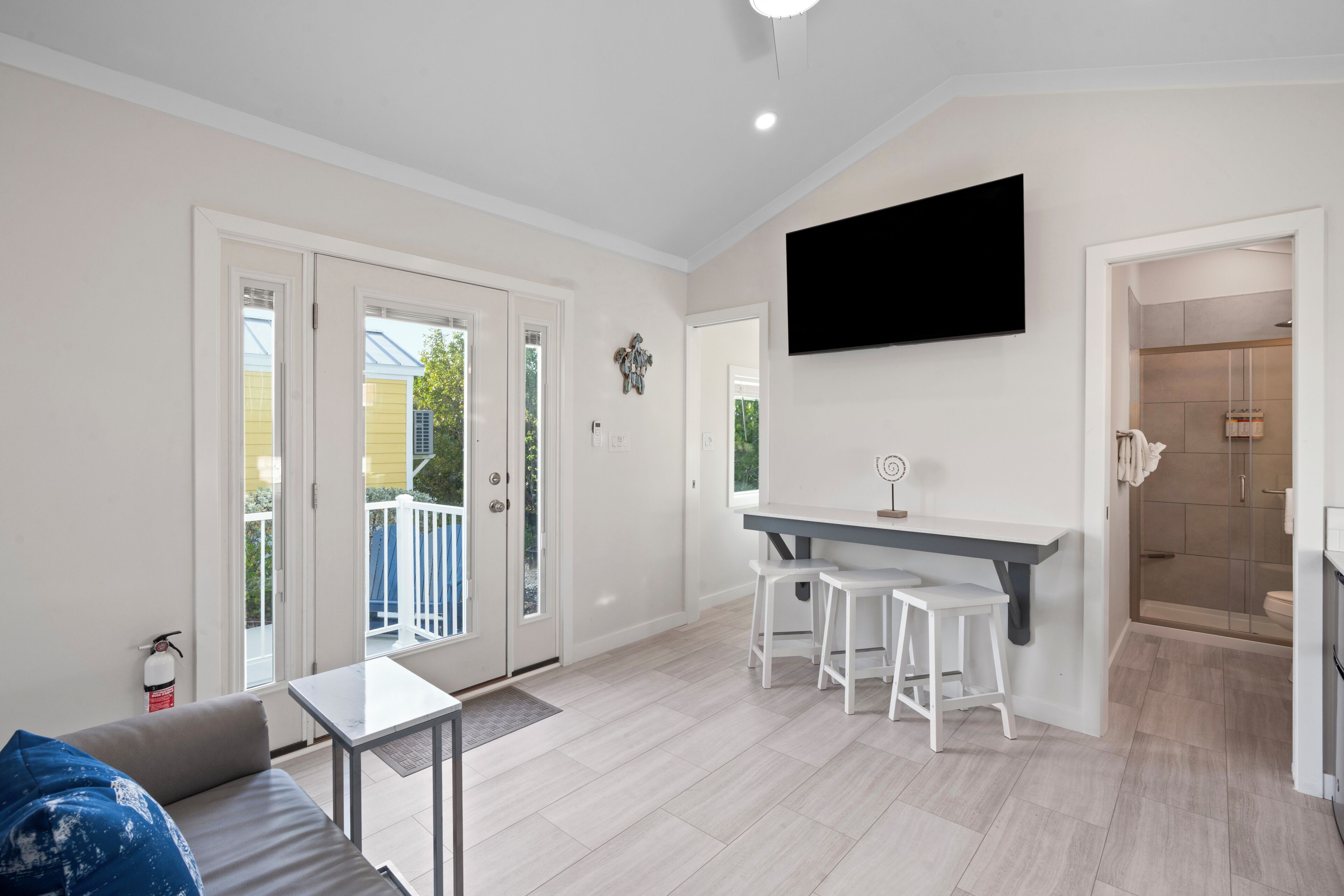 A modern living room with a sofa, a coffee table, a TV mounted on the wall, a kitchen area with a counter and stools, and a sliding glass door leading to an outdoor area.
