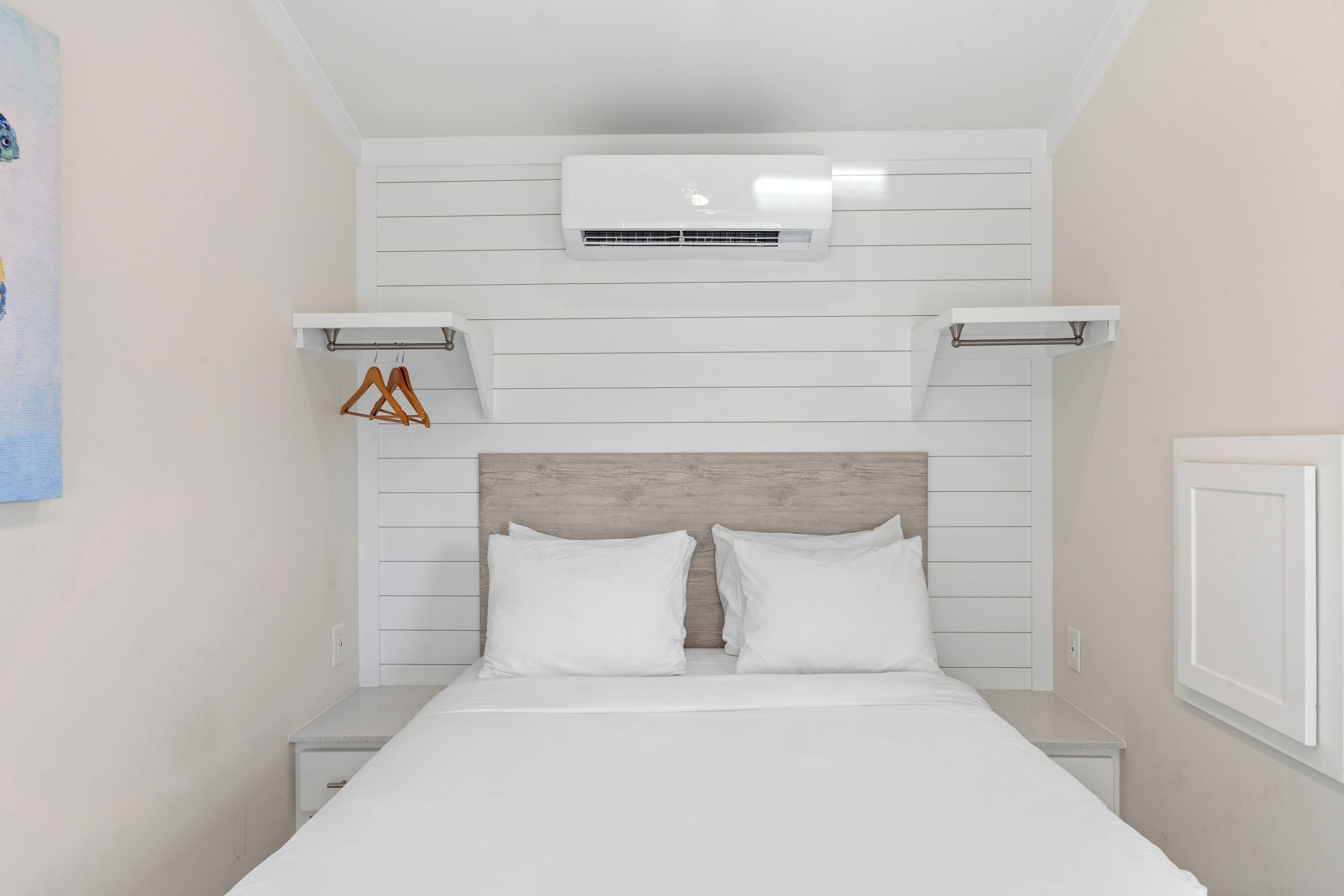 Ein Schlafzimmer mit Bett, weißen Wänden, einem hölzernen Kopfteil, einer Klimaanlage und einem kleinen Regal mit Kleiderbügeln.