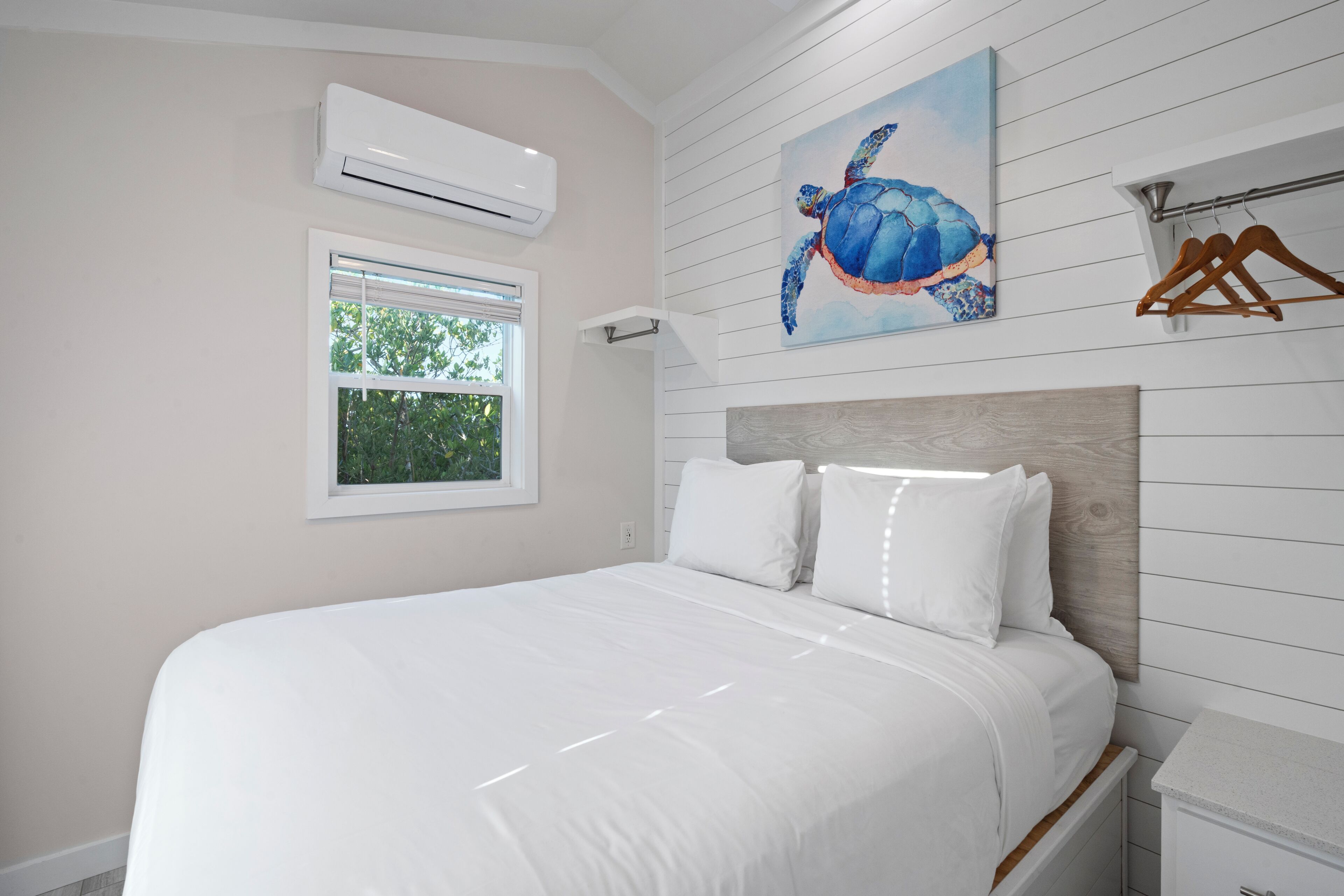 Ein Schlafzimmer mit Bett, weißen Wänden, einem Fenster, einer Klimaanlage, einem Holzkopfstück und einem Bild von einer Schildkröte.