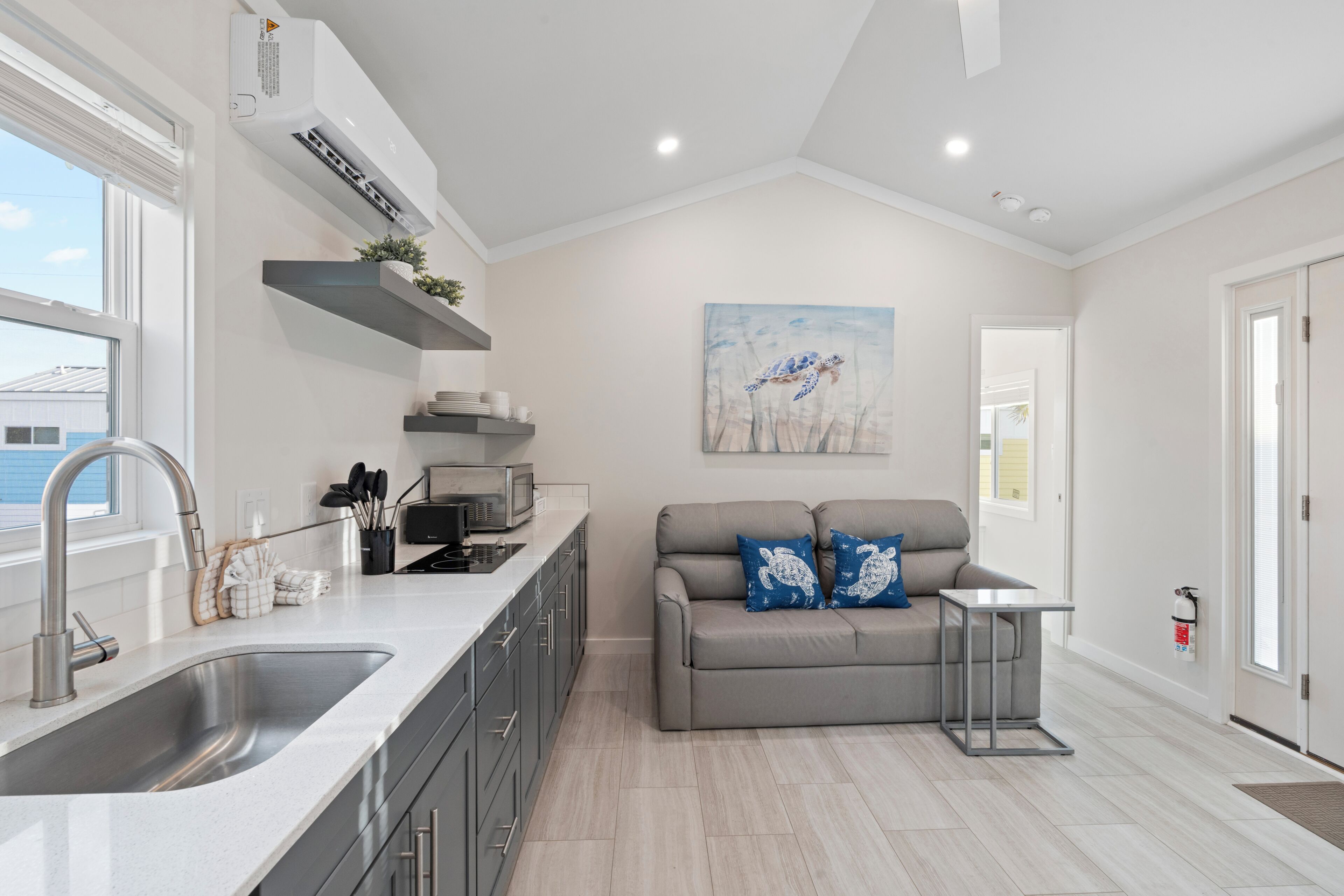 A modern kitchen with a built-in oven, a stainless steel sink, and a grey sofa with blue cushions.