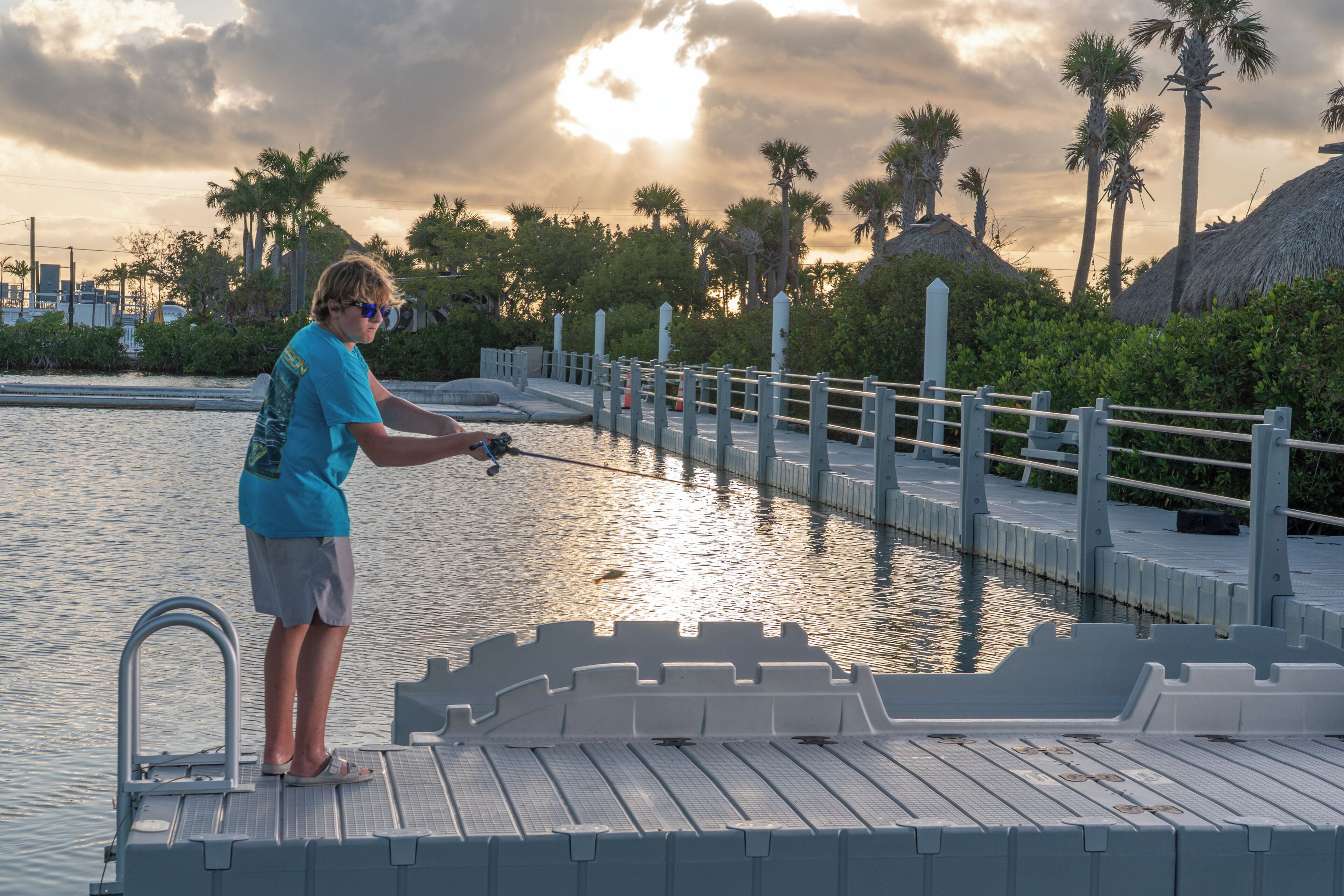 A person fishing on a dock with a railing and palm trees in the background.