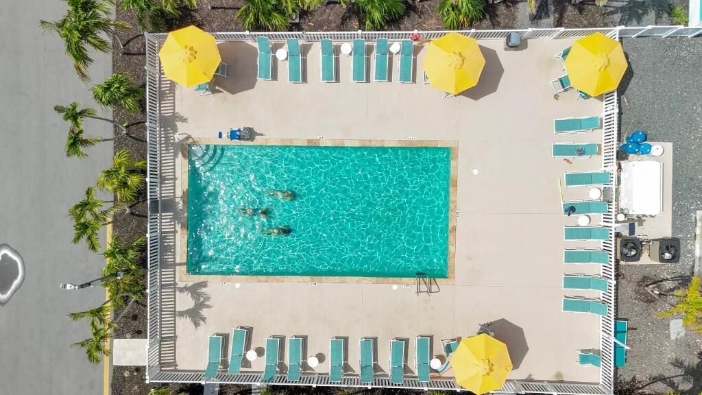 An aerial view of a rectangular swimming pool with clear blue water, surrounded by lounge chairs and yellow umbrellas.