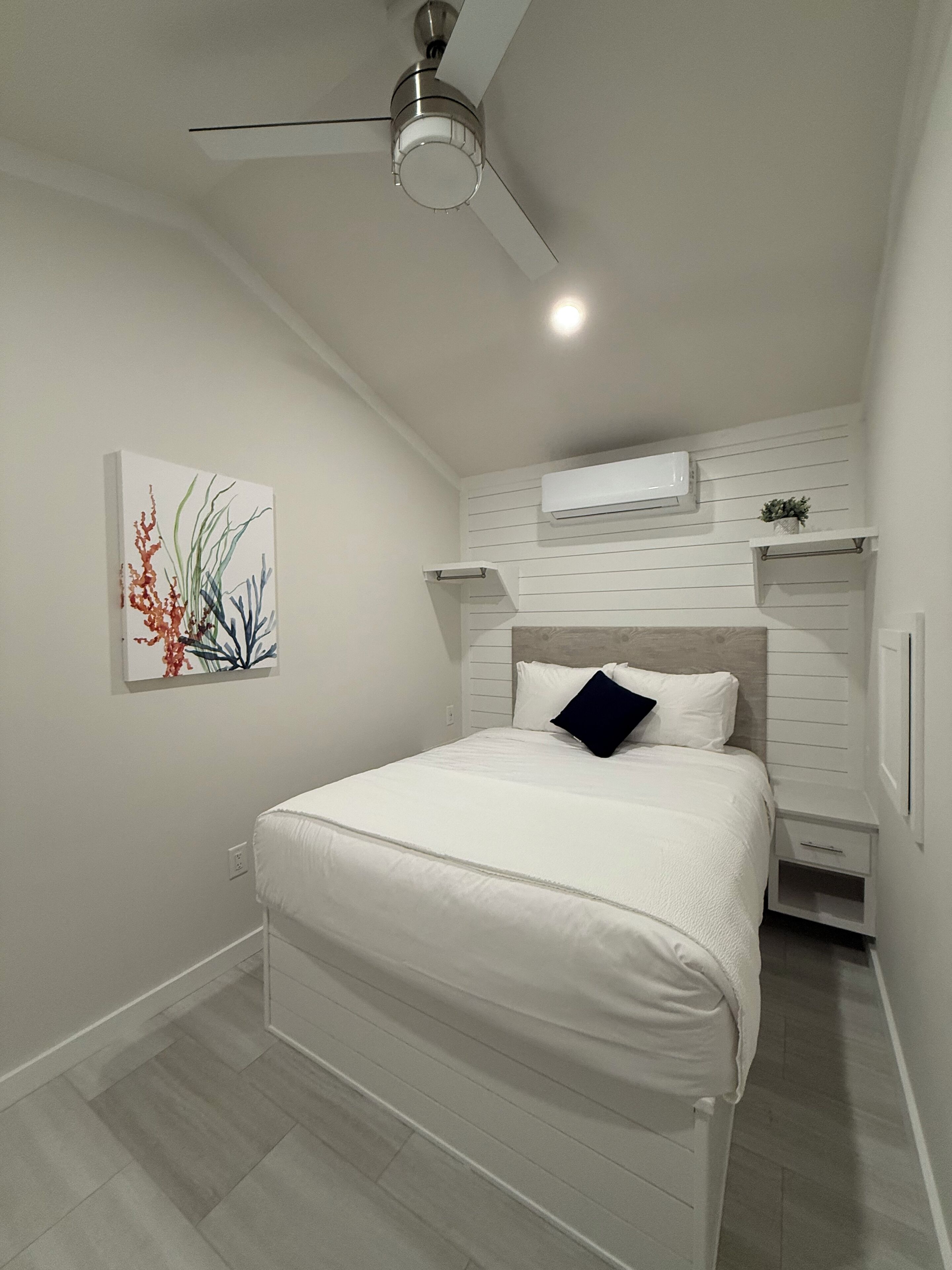 Een slaapkamer met een bed, een plafondventilator, een airconditioningapparaat, een nachtkastje en een wandplank.