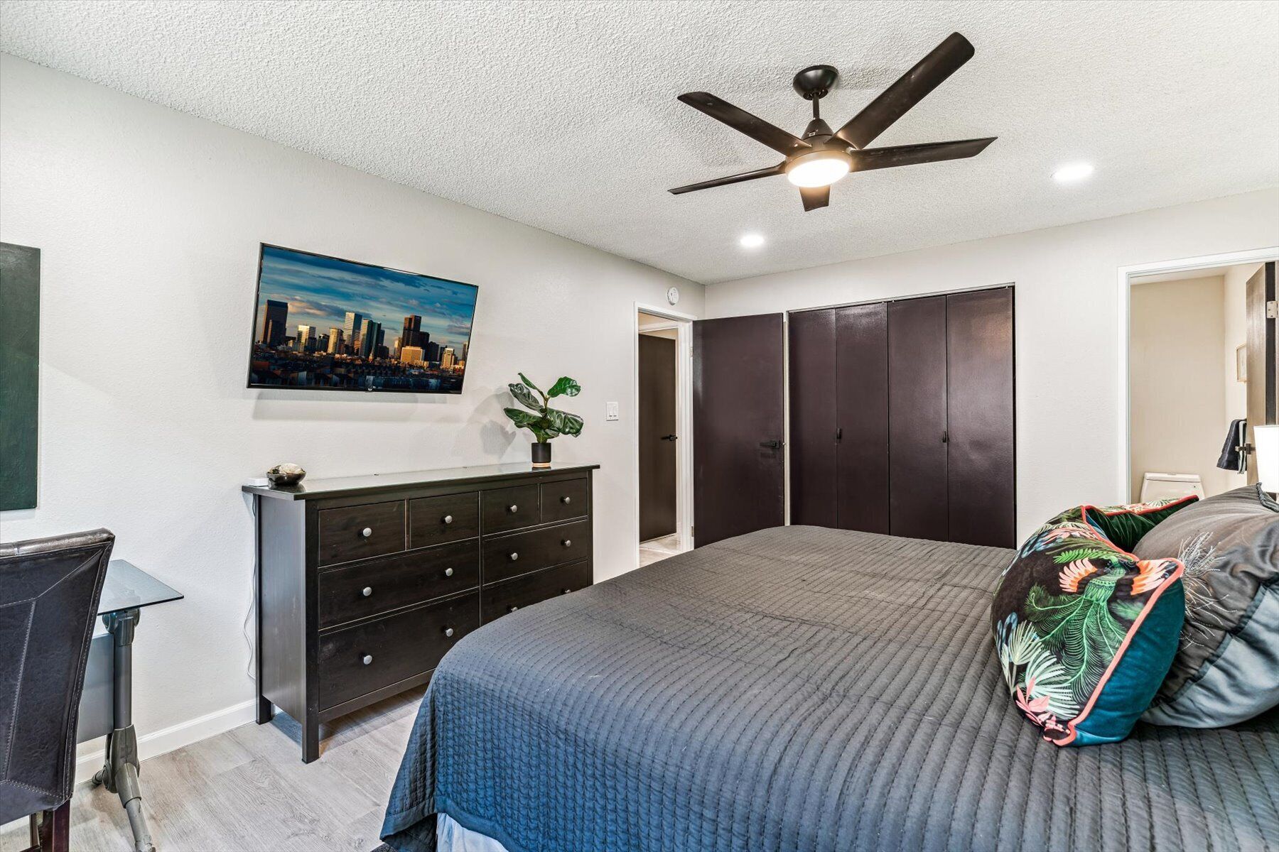 Une chambre à coucher comprenant un lit, une commode, un ventilateur de plafond et un tableau représentant un paysage urbain.